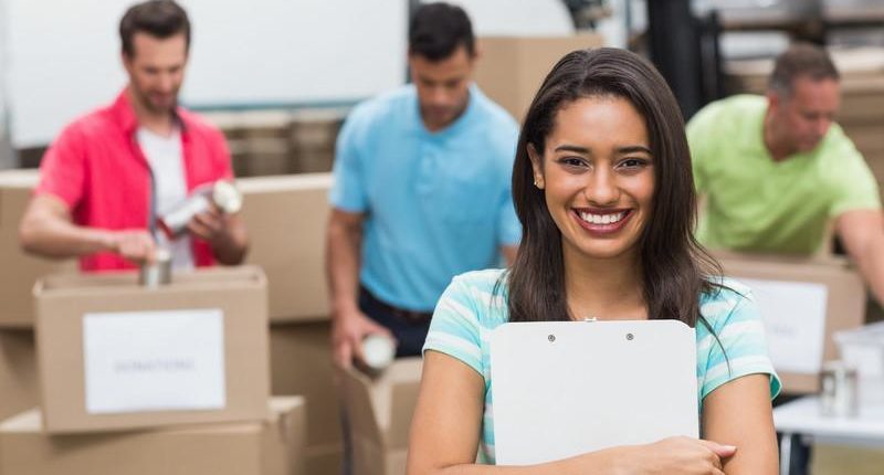 smiling woman holding a clipboard and behind her men are packing boxes