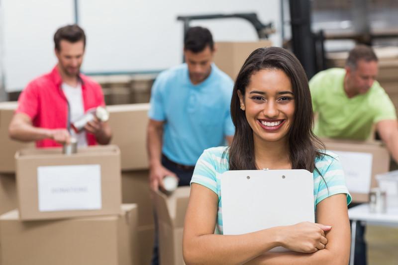 smiling woman holding a clipboard and behind her men are packing boxes