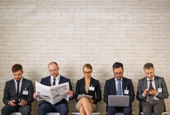 corporate employees seated in a row holiding phones, laptops and newspapers.