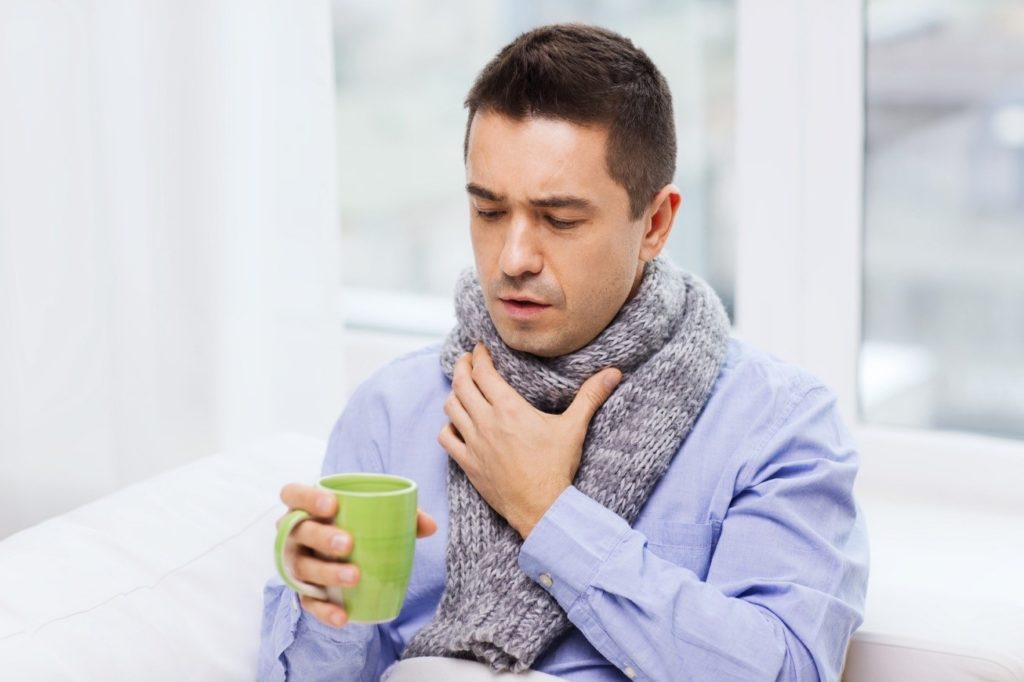 Man wearing scarf, clasping his throat, holding a mug