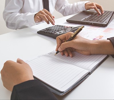 person writing into a notebook, person using a laptop and calculator