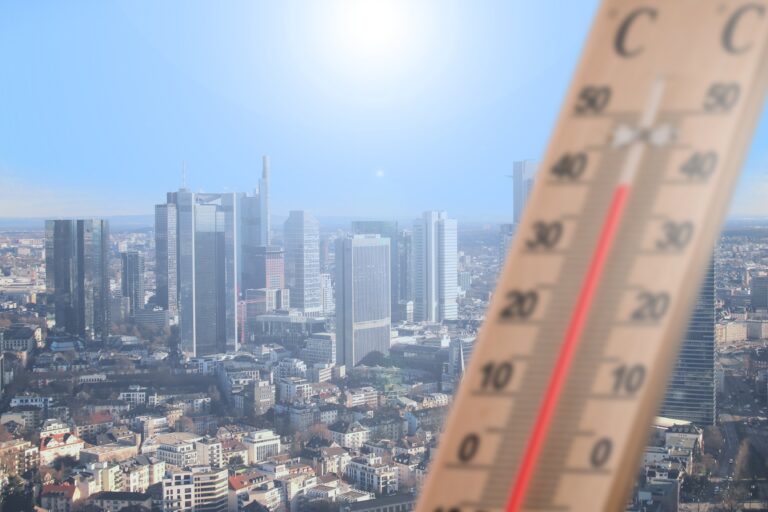 A city under a sunny sky in the background of the image, with a thermometre in the foreground