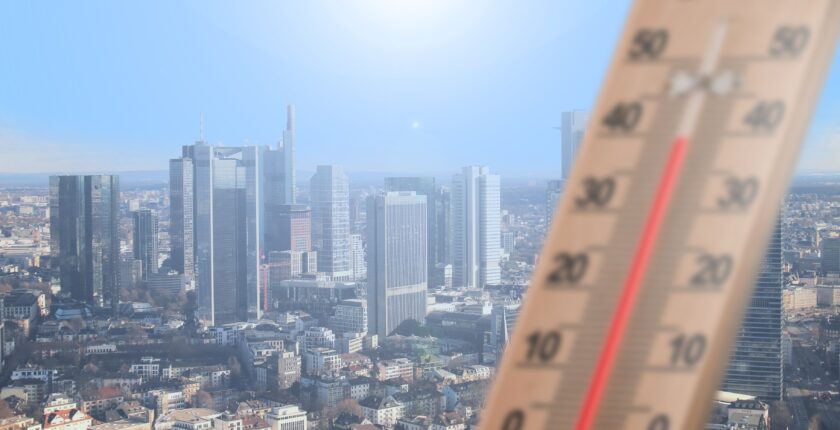 A city under a sunny sky in the background of the image, with a thermometre in the foreground
