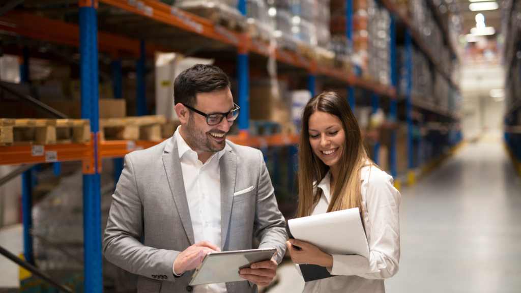 Two professionals reviewing documents and a tablet together in a warehouse, with industrial shelving and inventory in the background.