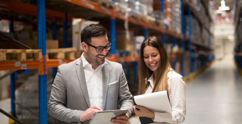 Two professionals reviewing documents and a tablet together in a warehouse, with industrial shelving and inventory in the background.