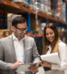 Two professionals reviewing documents and a tablet together in a warehouse, with industrial shelving and inventory in the background.