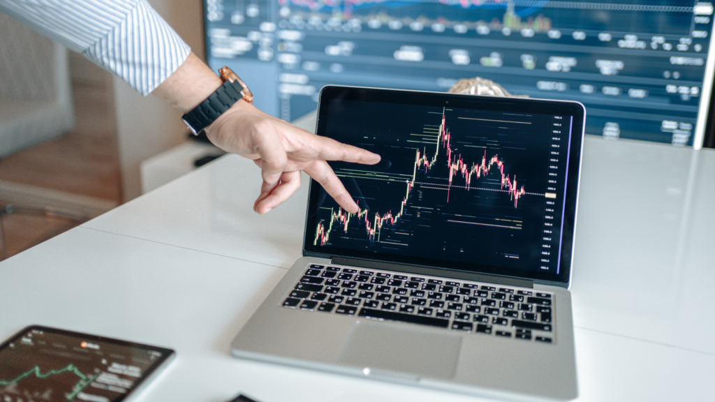 A person pointing to financial market trends displayed on a laptop screen, with additional stock market charts visible in the background.