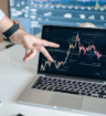 A person pointing to financial market trends displayed on a laptop screen, with additional stock market charts visible in the background.